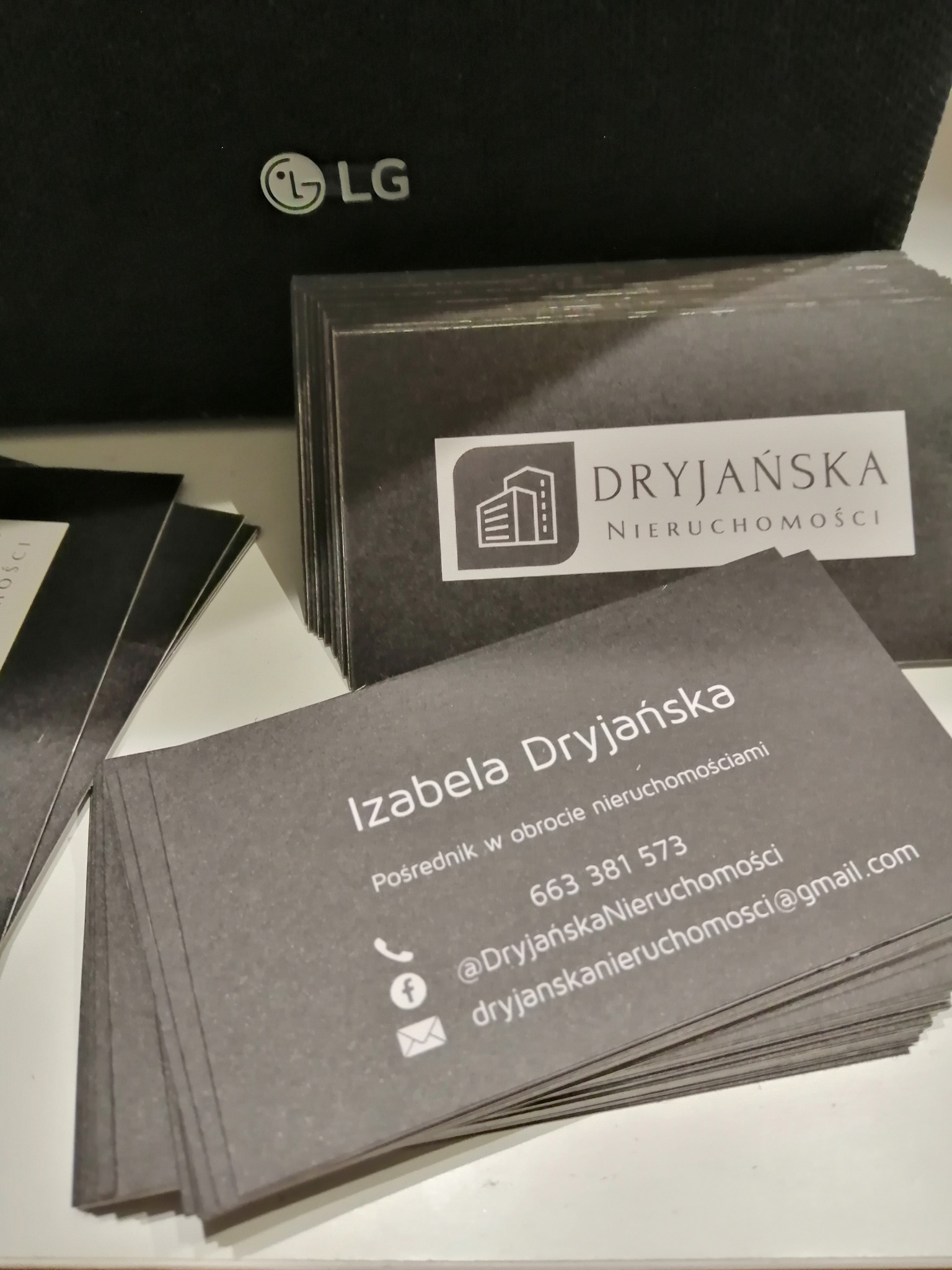 Stos wizytówek firmy Dryjańska Nieruchomości, z logo przedstawiającym budynek i danymi kontaktowymi pośrednika Izabeli Dryjańskiej, na tle czarnego pudełka z logo LG.