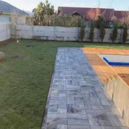 Świeżo założony trawnik z rolki w Ostrowie Wielkopolskim, z młodymi drzewkami i ścieżką z kostki prowadzącą do drewnianego tarasu przy basenie.