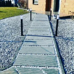Ścieżka z szarych płyt betonowych oddzielonych białym grysem, otoczona szarym i białym żwirem, oświetlona lampami ogrodowymi, prowadząca do domu z cegły.