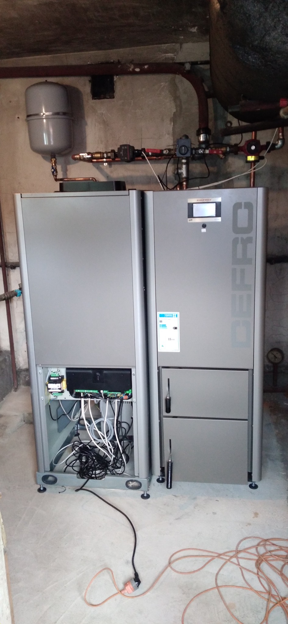 Szary, dwuczęściowy piec DEFRO z otwartym panelem bocznym, ukazującym wewnętrzne okablowanie i elektronikę, zamontowany w surowym pomieszczeniu z betonową podłogą i widocznymi instalacjami...