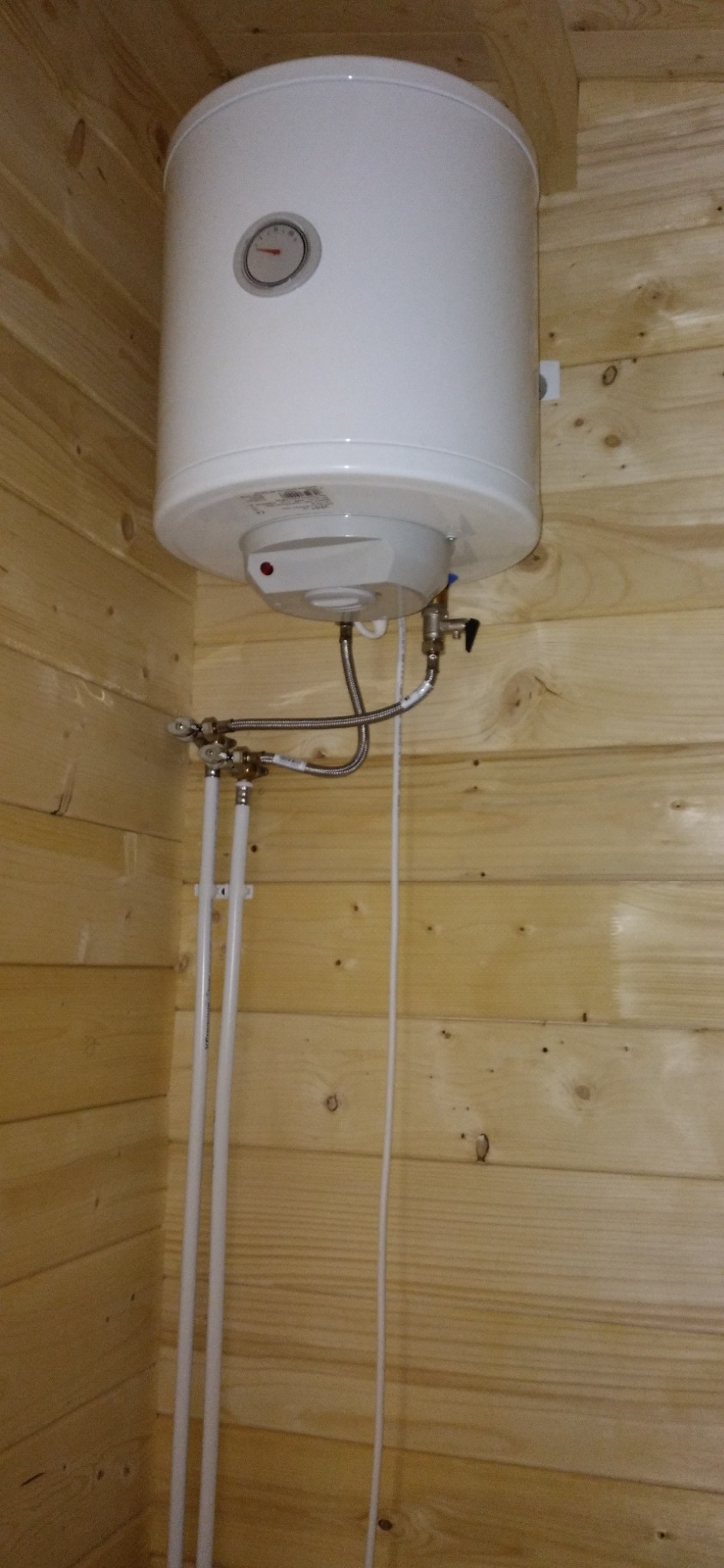 Bojler elektryczny zamontowany w rogu pomieszczenia wyłożonego boazerią, widoczne przyłącza hydrauliczne i przewody.