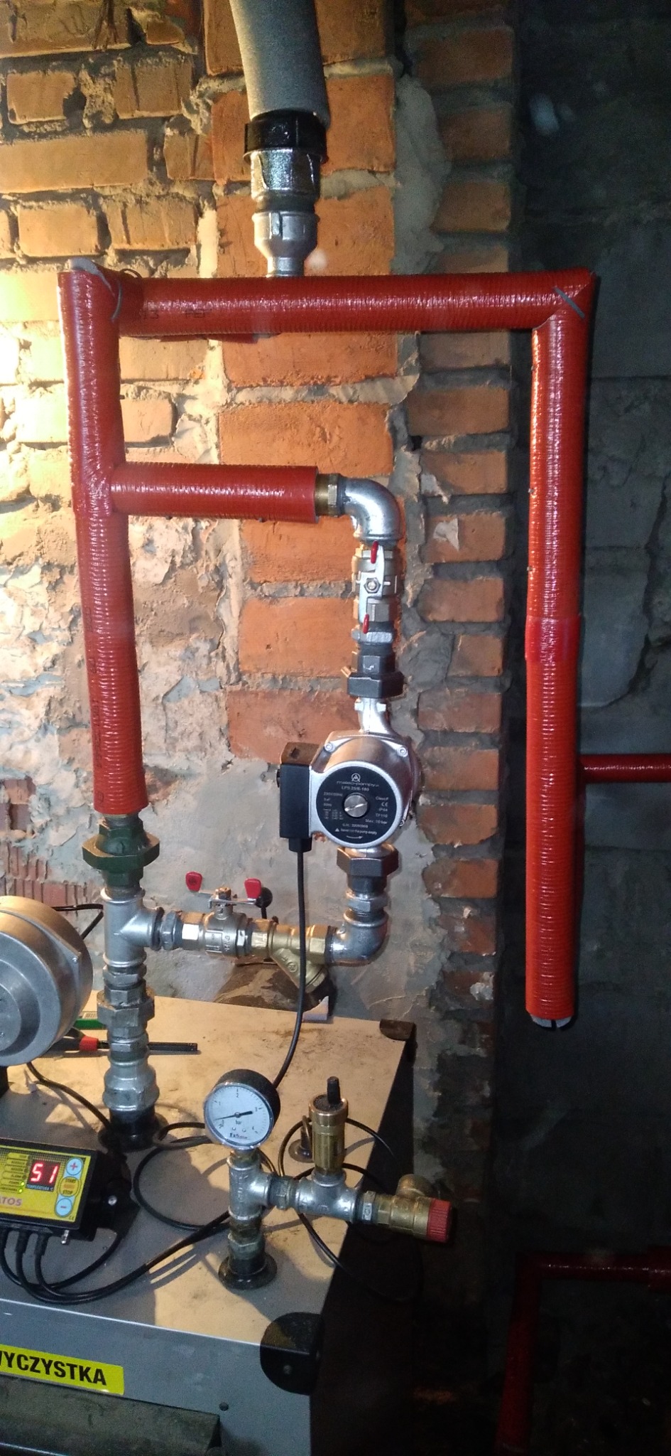 Fragment instalacji centralnego ogrzewania z pompą obiegową, rurami w czerwonej izolacji, manometrem i sterownikiem, zamontowany na tle ceglanej ściany.