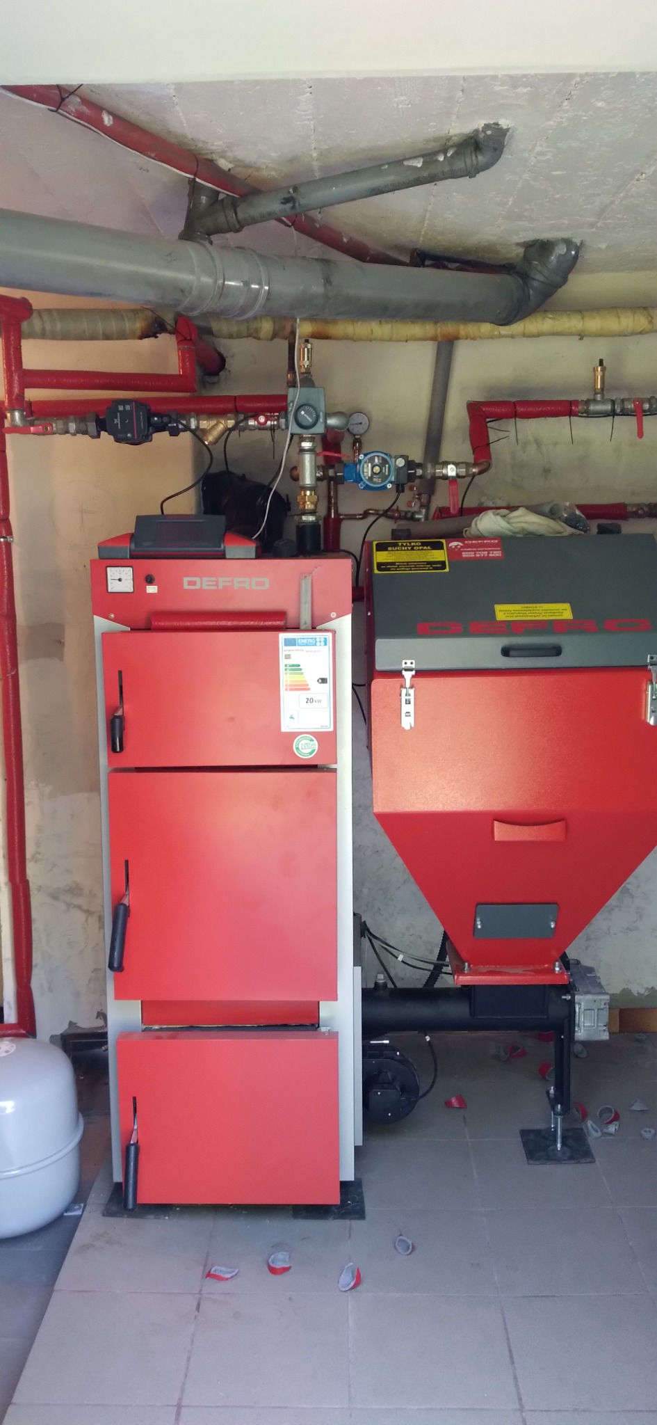 Czerwony piec Defro z podajnikiem na ekogroszek, zainstalowany w kotłowni, widoczne rury instalacji centralnego ogrzewania, na podłodze resztki opakowań po montażu.
