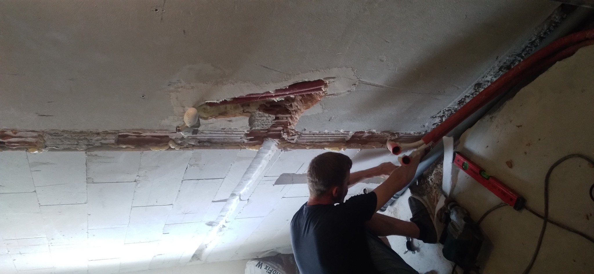 Hydraulik montuje rury pod sufitem, widoczne uszkodzenia w suficie i wystające rury, poziomica na ścianie.