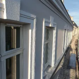 Odnowiona elewacja budynku z białymi, zdobionymi obramowaniami okien, widok z rusztowania w słoneczny dzień.