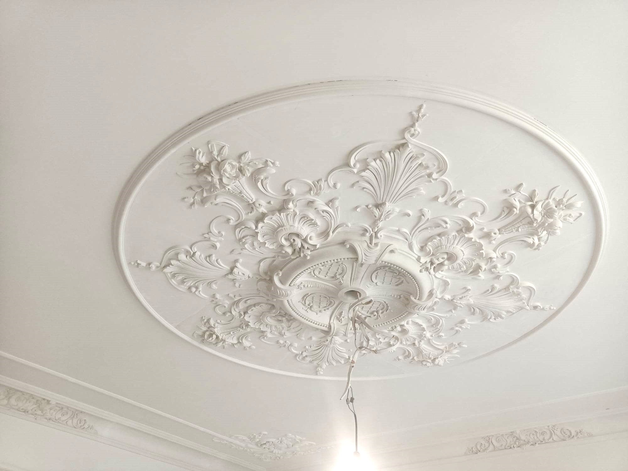 Biały sufit z okrągłą, ozdobną sztukaterią w stylu rokoko, z widocznymi kablami zwisającymi ze środka.