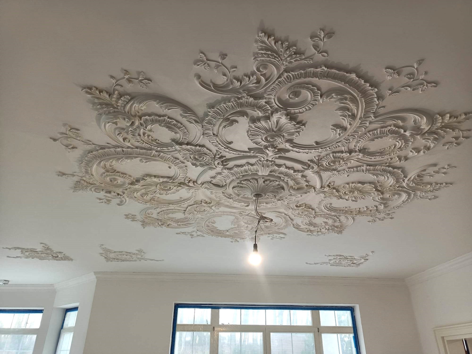 Biały sufit z ozdobnym, gipsowym ornamentem w stylu rokoko, oświetlony żarówką na kablu, widok z dołu.