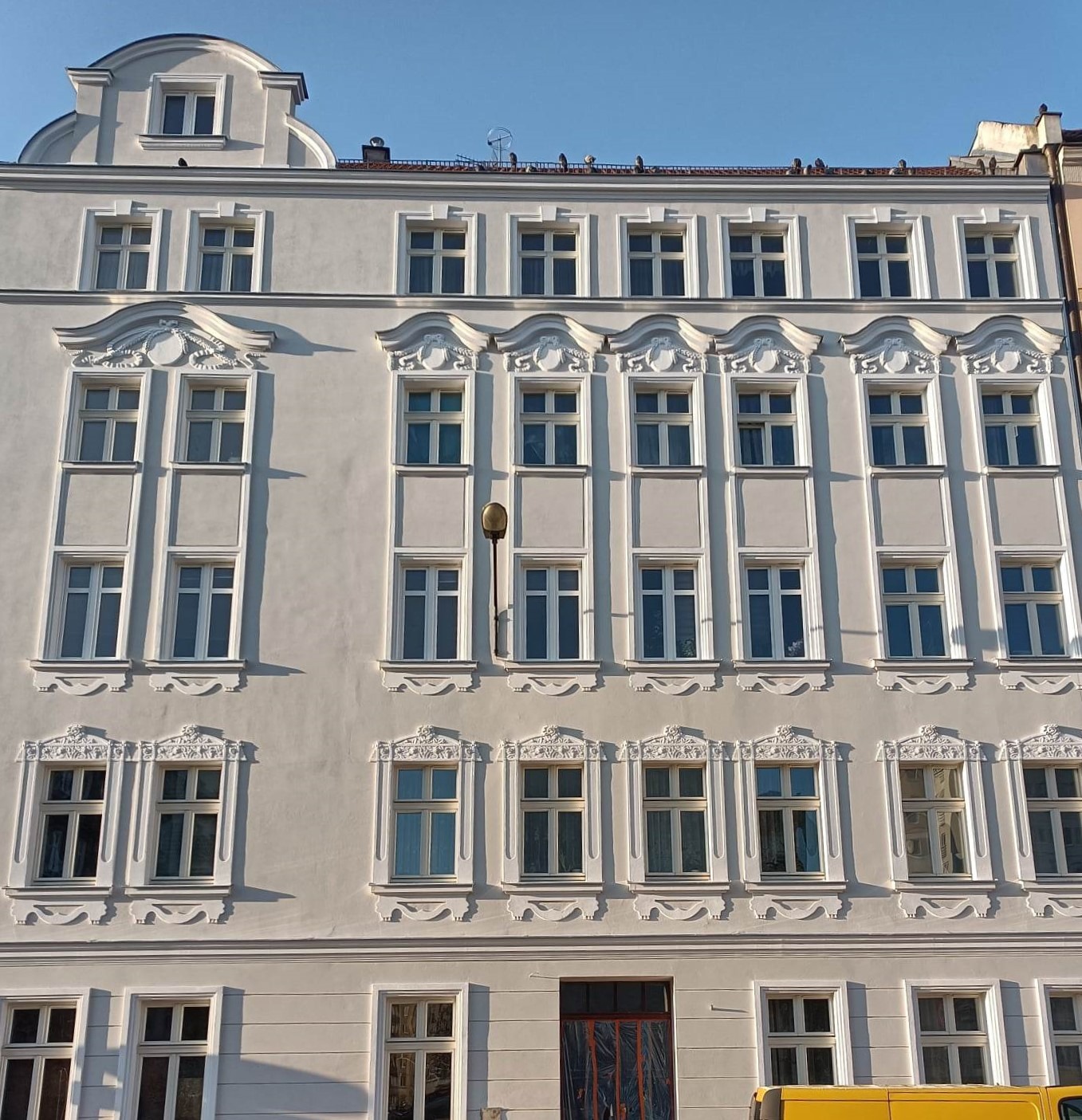 Fasada odnowionej, jasnoszarej kamienicy z licznymi oknami i dekoracyjnymi sztukateriami, oświetlona popołudniowym słońcem.