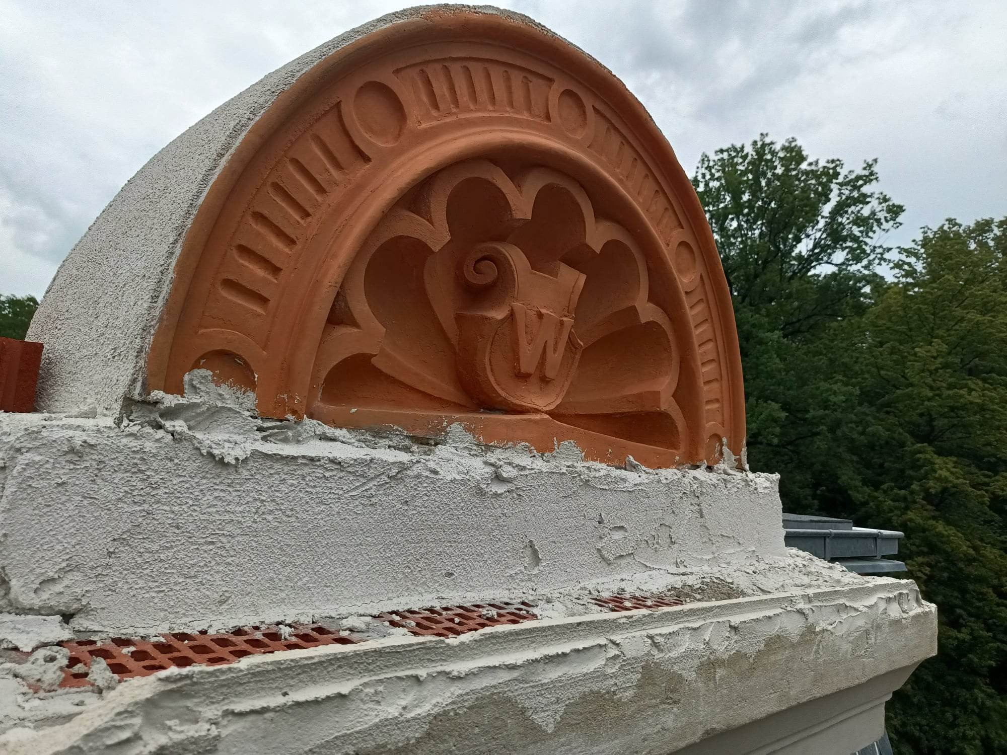 Ozdobny, terakotowy tympanon z monogramem litery 'W' na szczycie budynku, w trakcie prac murarskich, z widocznym świeżym tynkiem i kratką wentylacyjną pod spodem, na tle zieleni drzew i pochmurnego...