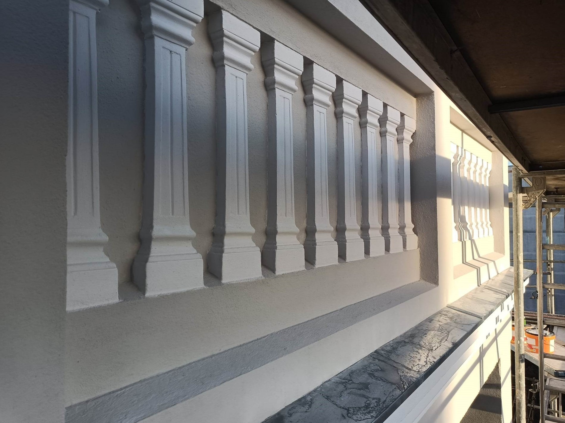 Odrestaurowana, pomalowana na biało balustrada balkonowa z kolumnami, widoczna z dołu, z fragmentem rusztowania po prawej stronie.