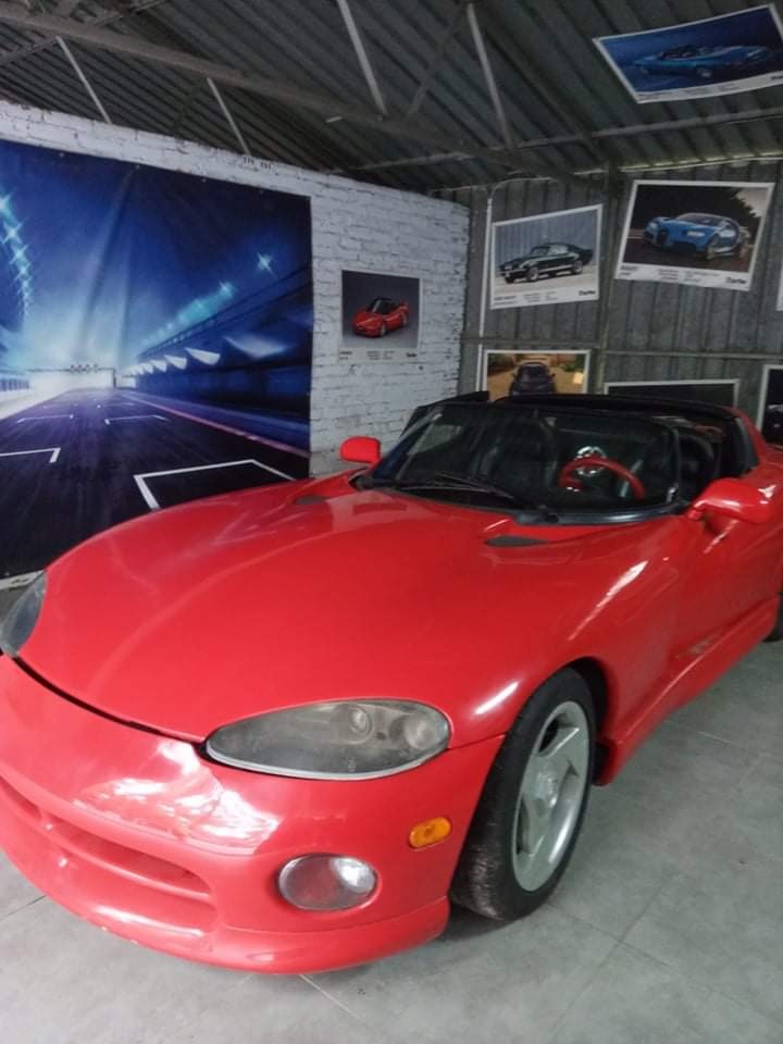 Czerwony sportowy kabriolet Dodge Viper w warsztacie z plakatem toru wyścigowego i zdjęciami innych samochodów na ścianie z białej cegły.