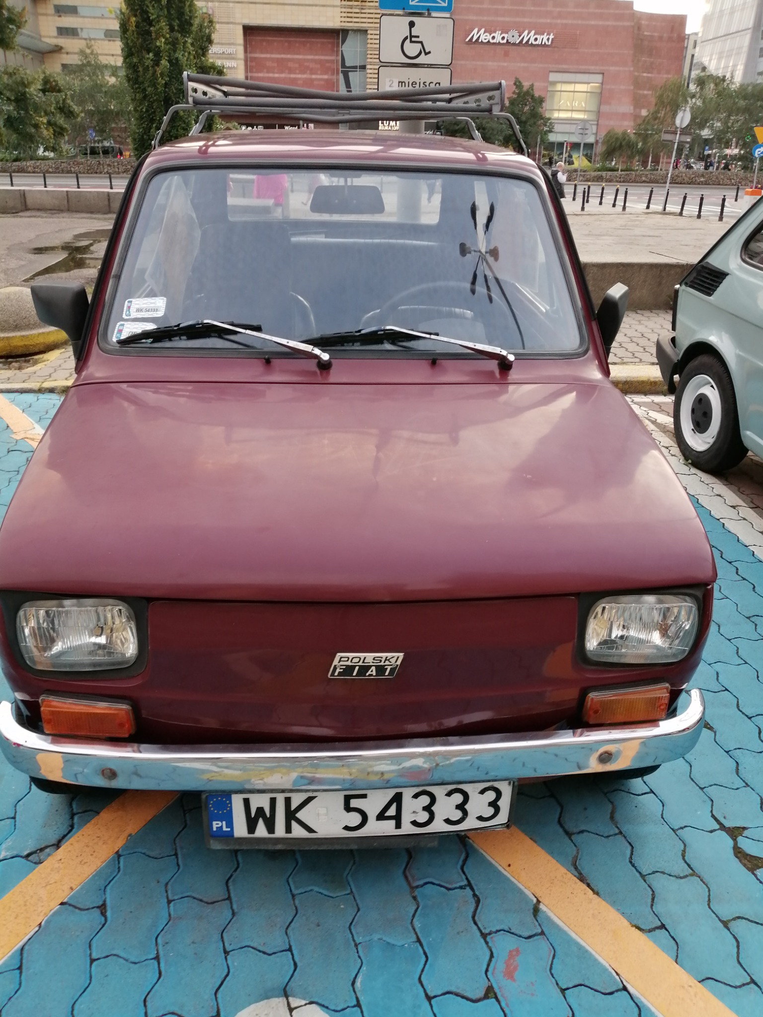 Przód bordowego Fiata 126p z bagażnikiem dachowym na parkingu z niebieską kostką i żółtymi liniami, w tle budynek Media Markt.
