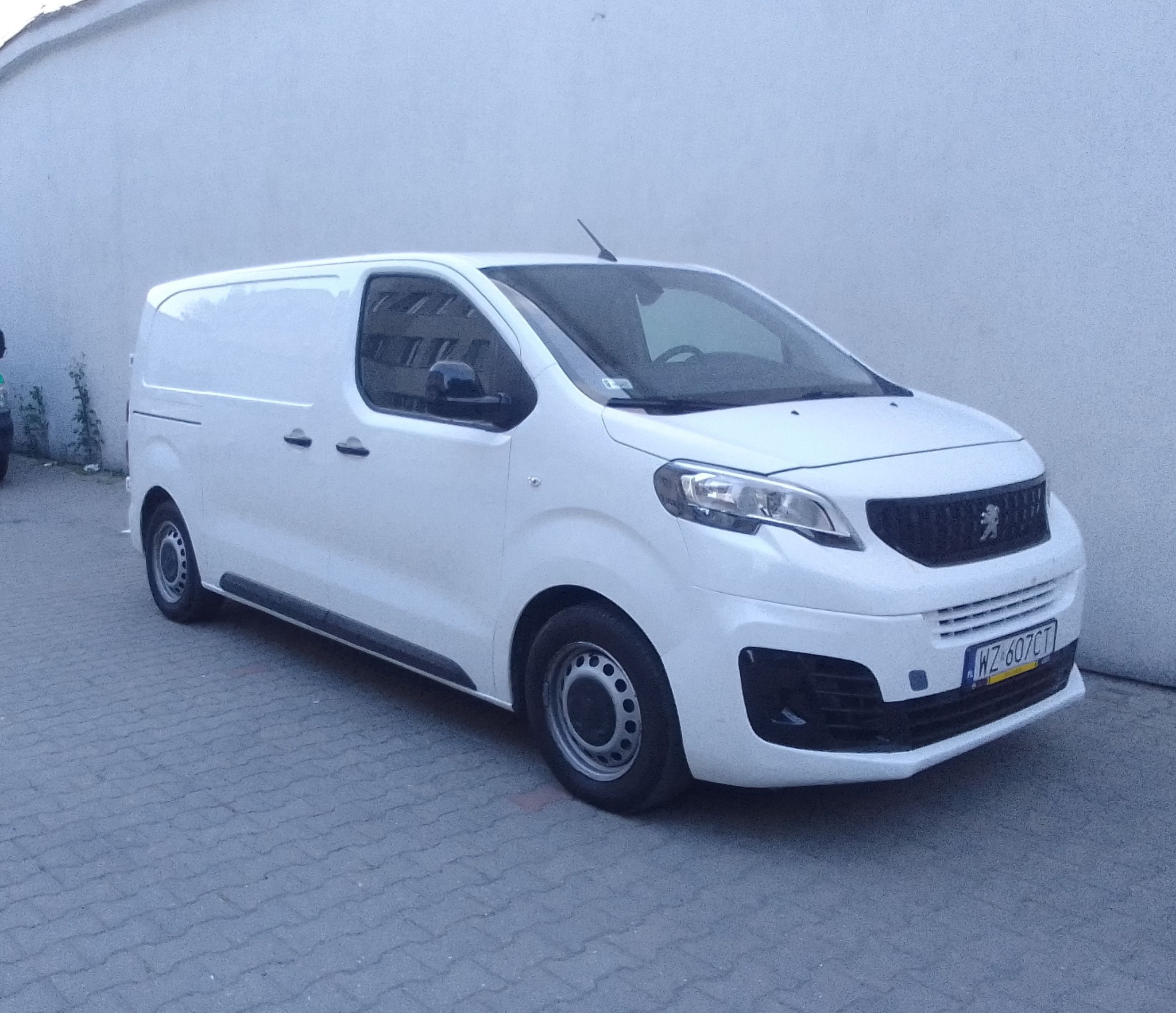 Biały samochód dostawczy marki Peugeot z widoczną tablicą rejestracyjną WZ 607CT zaparkowany na szarym chodniku na tle jasnej ściany budynku.