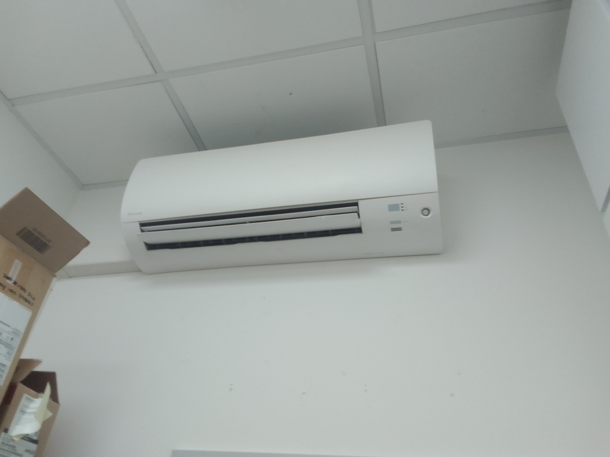 Biała jednostka klimatyzacji zamontowana na ścianie, widoczna spod sufitu kasetonowego, z kartonowym pudłem w tle po lewej stronie.