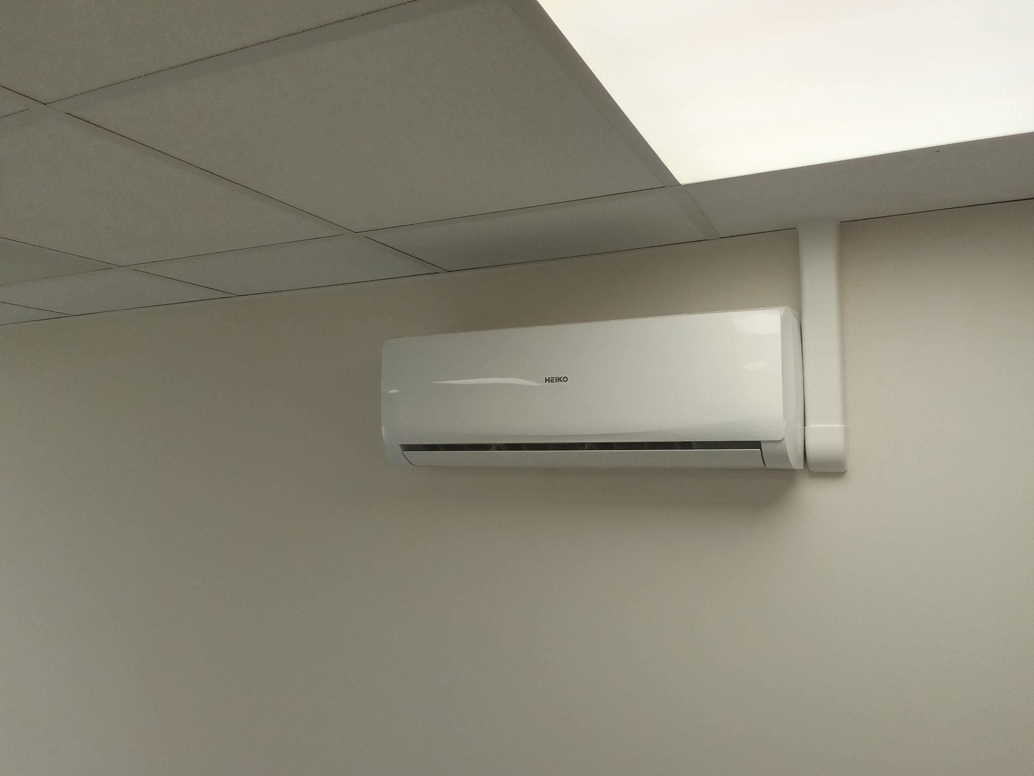 Biała jednostka klimatyzacji HEIKO zamontowana na jasnej ścianie w biurze, widoczne rury instalacyjne.