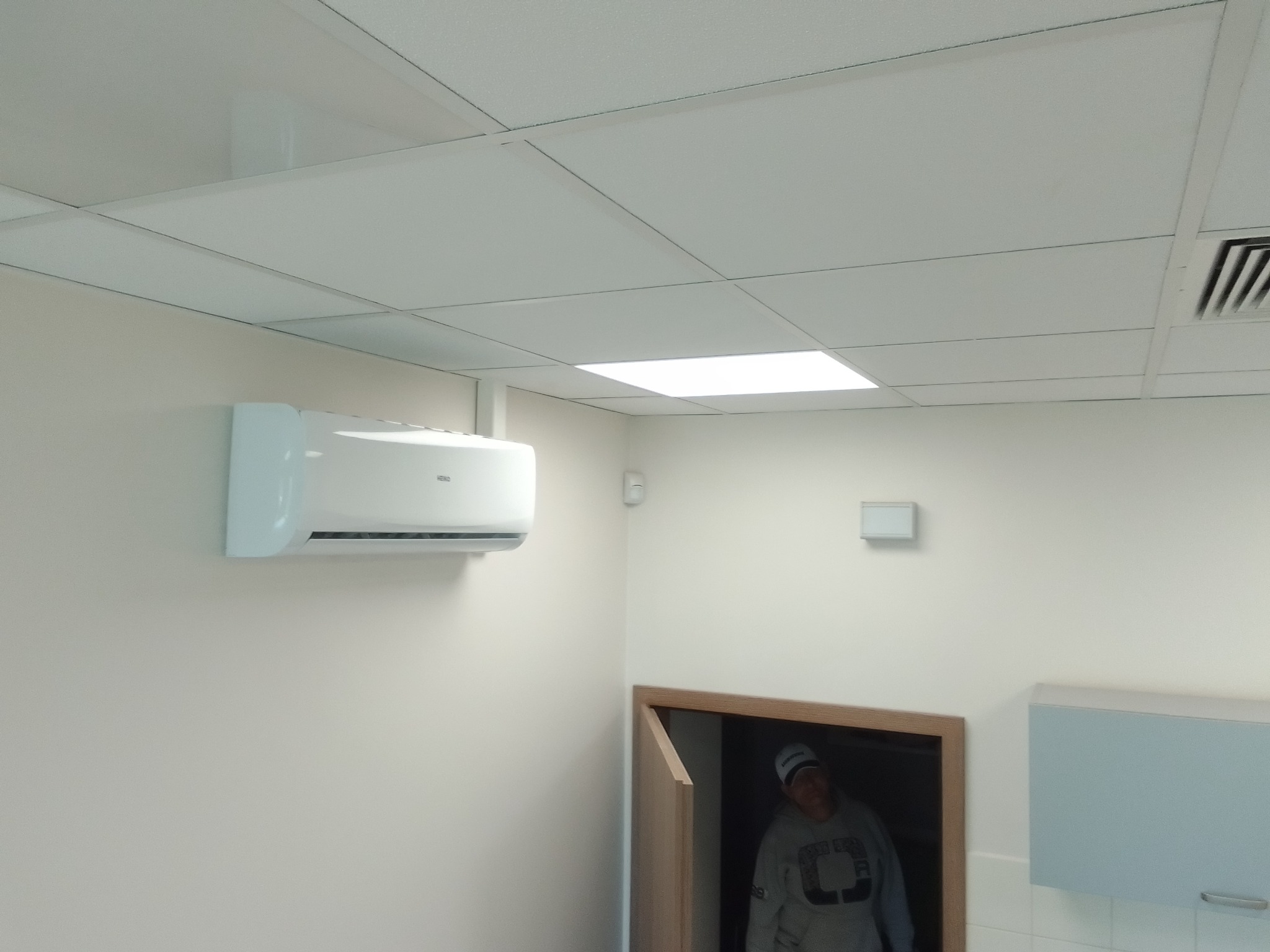 Biały klimatyzator ścienny zamontowany w pomieszczeniu z podwieszanym sufitem kasetonowym, widoczny fragment wentylacji i osoba w drzwiach w tle.