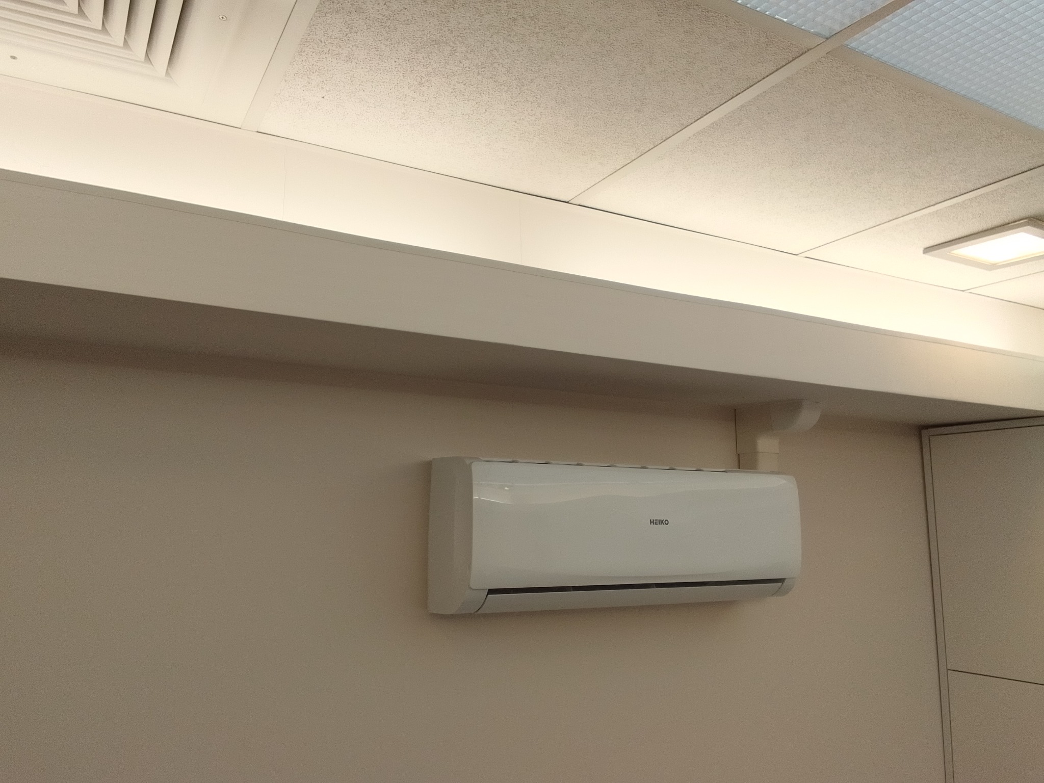 Biała jednostka klimatyzacji marki Heiko zamontowana na beżowej ścianie w biurze, widoczne rury instalacyjne i sufit kasetonowy z oświetleniem LED.