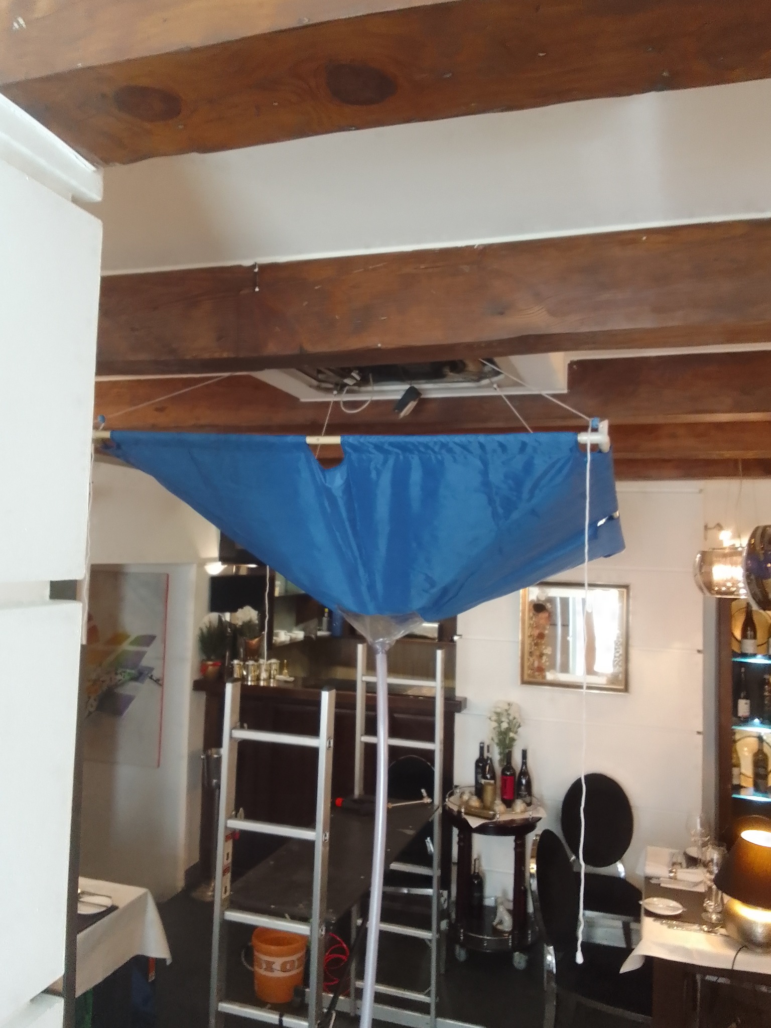 Montaż klimatyzacji w restauracji, widoczna niebieska plandeka zabezpieczająca przed kurzem, drabina i elementy instalacji pod sufitem.