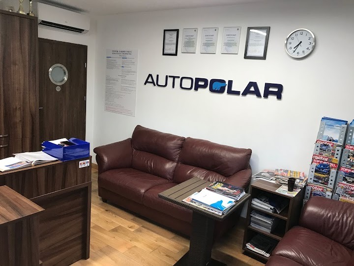 Wnętrze biura z brązową sofą, stolikiem z magazynami, regałem na prasę, logo firmy AUTOPOLAR na ścianie, klimatyzatorem ściennym i certyfikatami w ramkach.