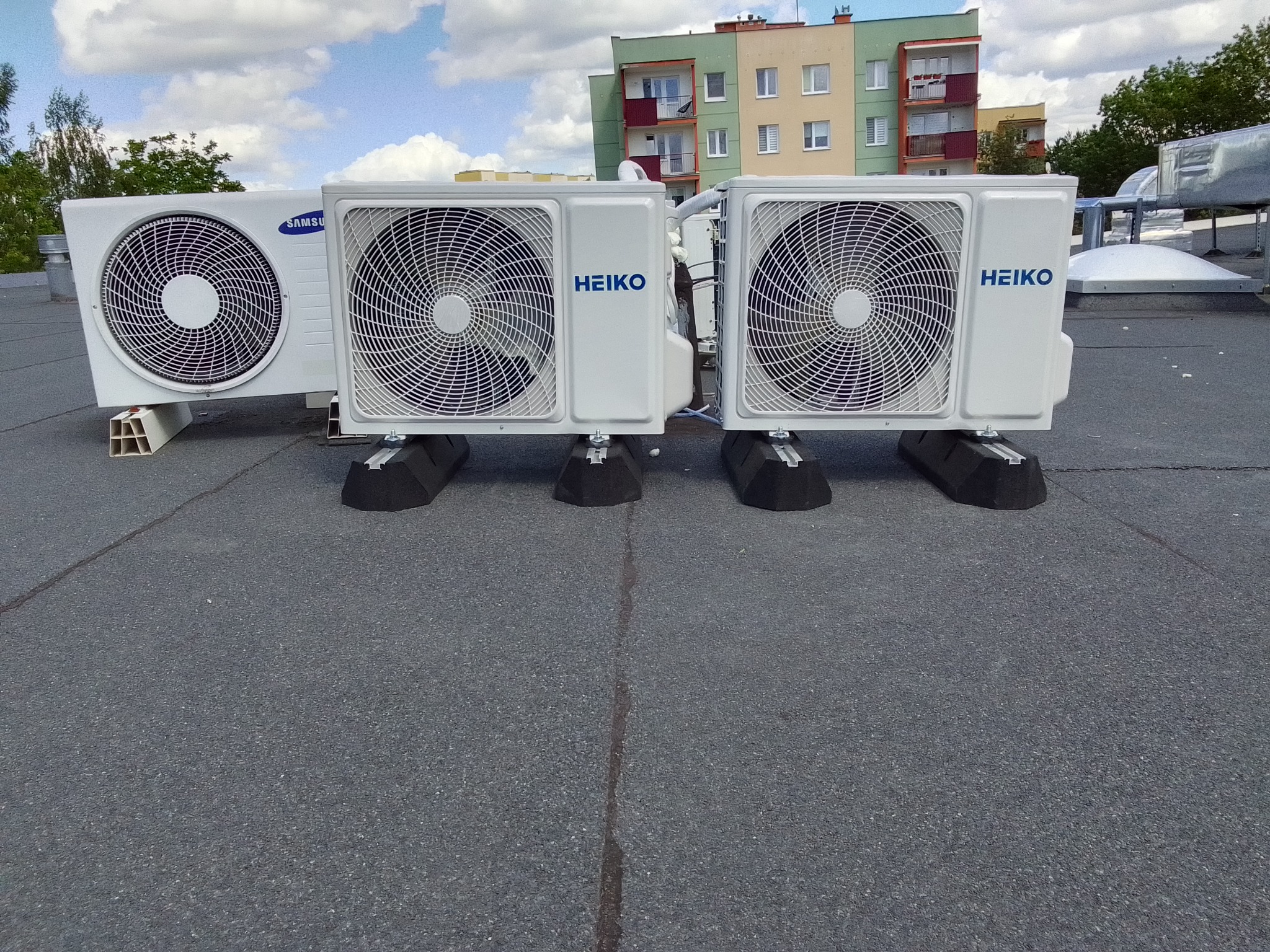 Trzy jednostki zewnętrzne klimatyzacji (Samsung i Heiko) zamontowane na płaskim dachu budynku mieszkalnego, widoczne przewody i wsporniki antywibracyjne.