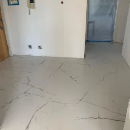 Białe, błyszczące płytki podłogowe z szarym wzorem marmuru w nowym mieszkaniu, widoczne gniazdka elektryczne, domofon i zabezpieczone folią drzwi.