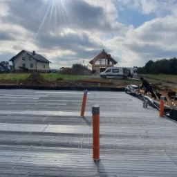 Przygotowanie podłoża pod budowę, widoczne rury wentylacyjne, czarna folia izolacyjna, w tle dom w trakcie budowy i dwa samochody dostawcze.