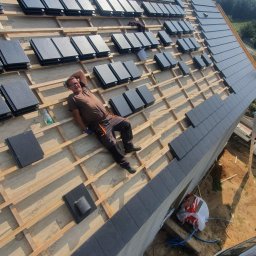 Zbudujecidom Spółka Cywilna - Pracownik relaksuje się na drewnianej konstrukcji dachu w trakcie układania ciemnoszarej dachówki ceramicznej, widoczne elementy mocujące i częściowo pokryty dach, butelka z napojem obok.