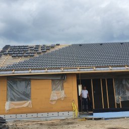 Zbudujecidom Spółka Cywilna - Budowa domu w technologii szkieletowej z częściowo ułożonym pokryciem dachowym z czarnej dachówki, dwóch pracowników na budowie, w tle widoczne panele słoneczne i pochmurne niebo.