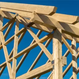 Konstrukcja drewniana dachu domu w trakcie budowy, widok od dołu na tle błękitnego nieba, widoczne metalowe łączniki.