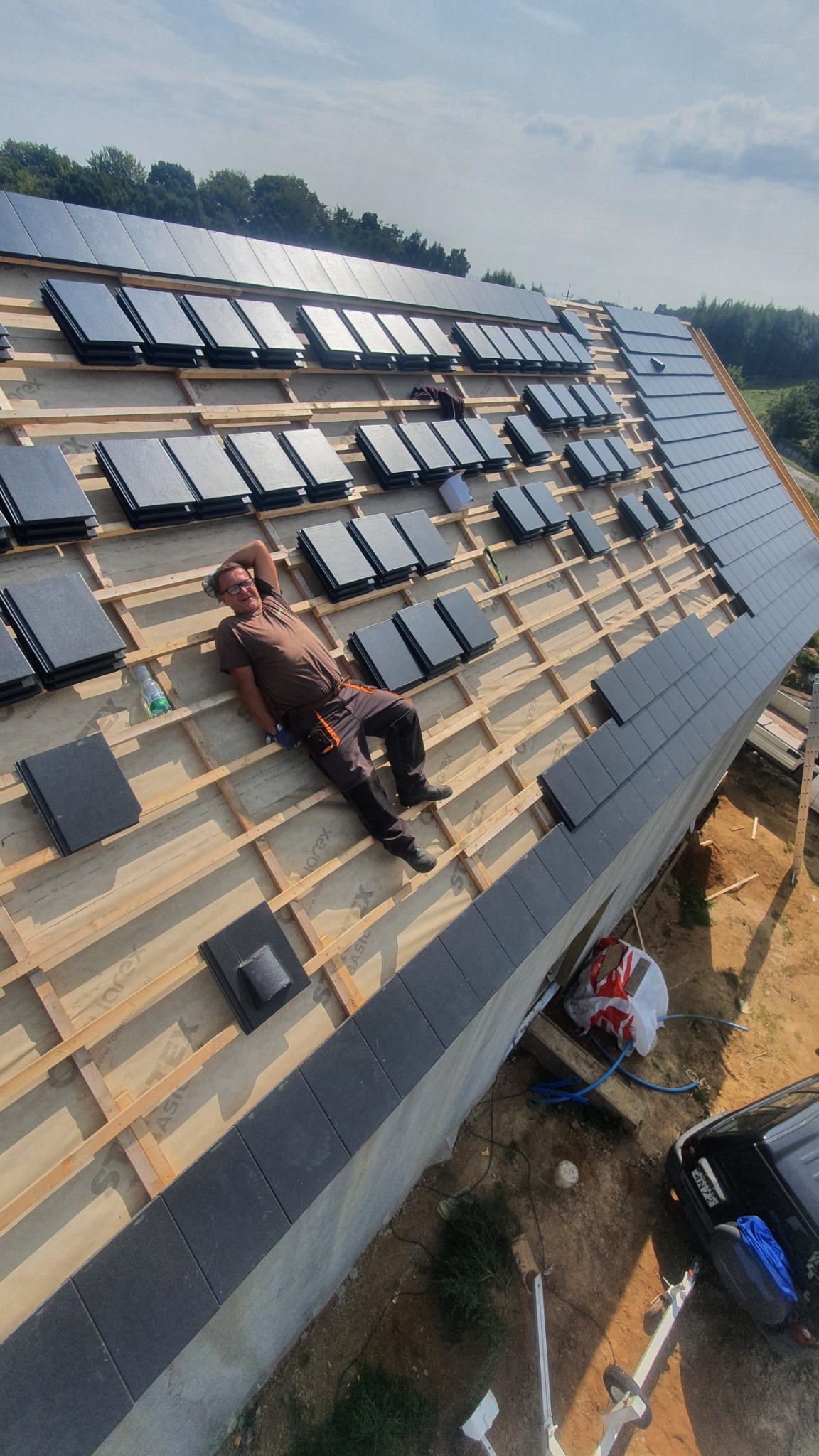 Pracownik relaksuje się na drewnianej konstrukcji dachu w trakcie układania ciemnoszarej dachówki ceramicznej, widoczne elementy mocujące i częściowo pokryty dach, butelka z napojem obok.