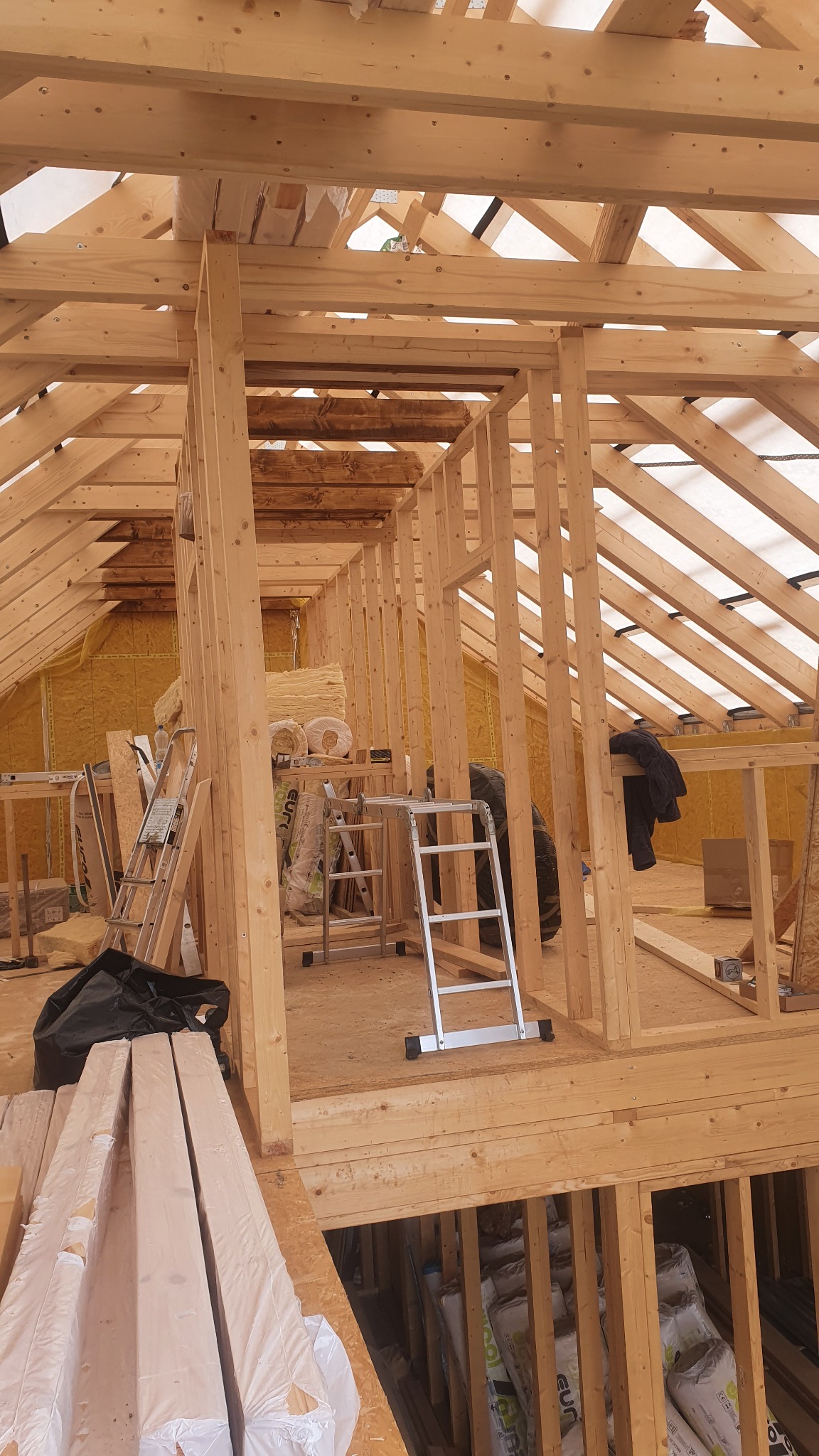 Wnętrze domu szkieletowego w trakcie budowy, widoczne drewniane elementy konstrukcyjne, izolacja, drabiny i materiały budowlane