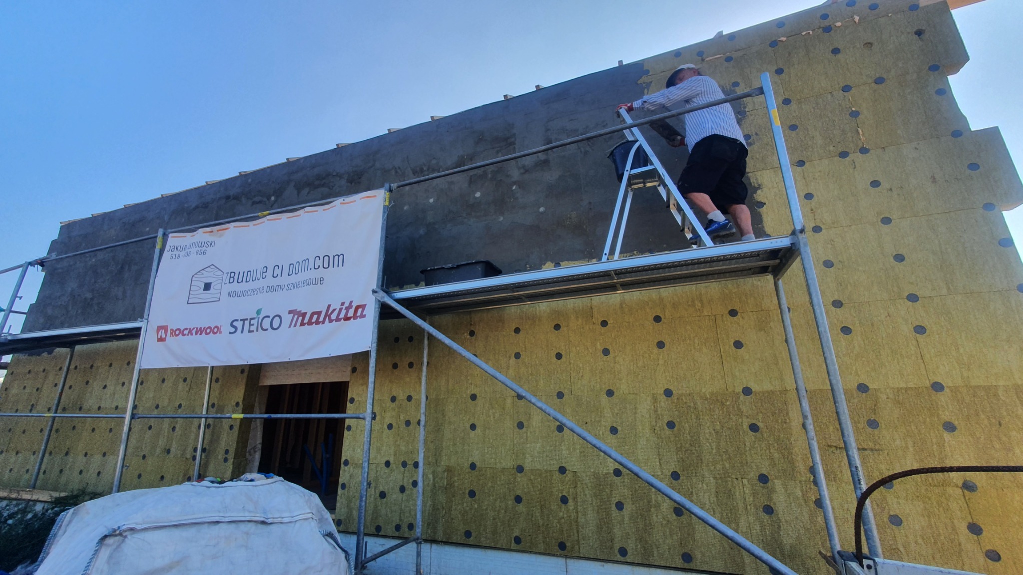 Monolityczna ściana budynku w trakcie ocieplania wełną mineralną, widoczny pracownik na rusztowaniu i baner reklamowy producenta materiałów budowlanych.