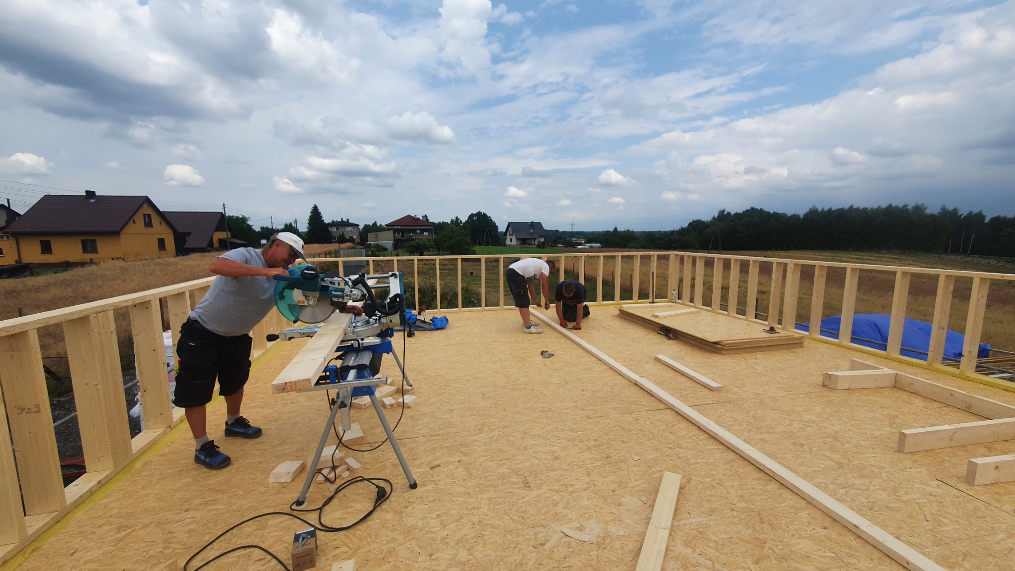 Budowa konstrukcji domu: stolarz tnie drewno piłą elektryczną na tle drewnianego szkieletu i krajobrazu wiejskiego.