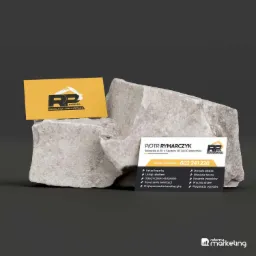 Dwie wizytówki firmy budowlanej (usługi mini koparką) leżące na szarym kamieniu. Jedna wizytówka jest żółta z czarnym logo, druga biała z czarnym tekstem i logo. Na wizytówkach widoczne usługi...