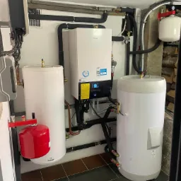 Instalacja grzewcza z białym kotłem Daikin, dwoma białymi zasobnikami wody i czerwonym naczyniem przeponowym Galmet, widoczne czarne rury izolowane i miedziane rurki, na podłodze ciemne płytki.