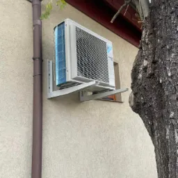Zewnętrzna jednostka klimatyzatora marki Daikin zamontowana na ścianie budynku, widoczna rynna spustowa i pień drzewa. Widok z dołu.