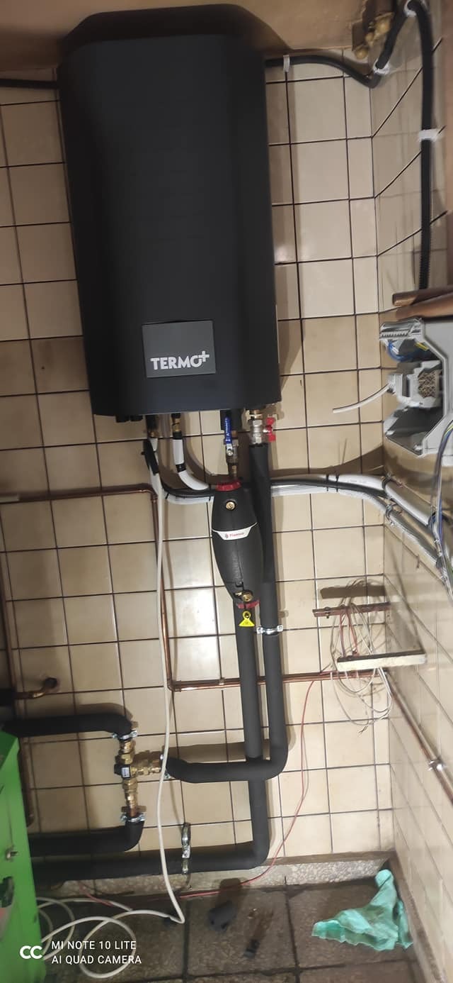 Czarny zasobnik ciepłej wody użytkowej marki TERMO+ z podłączonymi rurami w pomieszczeniu z wyłożonymi kafelkami ścianami.