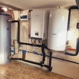 Instalacja kotłowni w piwnicy z szarym zbiornikiem akumulacyjnym, białym kotłem gazowym Kaisai, białym zasobnikiem ciepłej wody, pompą obiegową, filtrami i rurami w izolacji.