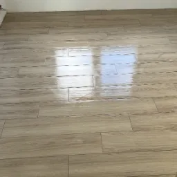 Ułożona jasna podłoga z płytek imitujących drewno, widoczne odbicie światła, fragment surowej ściany z cegły w rogu.