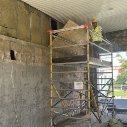 Tomasz Onyśko - Pracownik w kasku i kamizelce na rusztowaniu, montuje płytę. Widoczna surowa ściana z izolacją i instalacjami. Prace wykończeniowe w budynku.