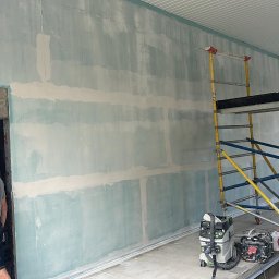 Tomasz Onyśko - Ściana w trakcie szpachlowania z widocznym rusztowaniem i pracownikiem w tle. Widoczny agregat szpachlarski. Prace wykończeniowe w jasnym pomieszczeniu.