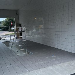 Tomasz Onyśko - Wyłożona szarymi płytkami podłoga i białą glazurą ściana w przestrzeni z otwartym przejściem. Widoczne rusztowanie i materiały budowlane. Prace w toku.
