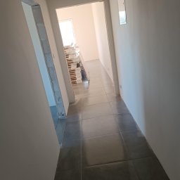 Tomasz Onyśko - Wykończony korytarz z szarymi płytkami podłogowymi. Widoczne przygotowane do montażu ościeżnice i sterta materiałów budowlanych w głębi. Świeżo pomalowane ściany.