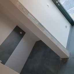 Tomasz Onyśko - Wnętrze w trakcie wykańczania: szara płytka na ścianie, biała ściana z oknem i belka z nałożoną gładzią, widoczne ślady po szpachlowaniu.