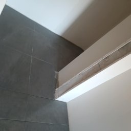 Tomasz Onyśko - Ciemnoszare, prostokątne płytki na ścianie, biały sufit i fragment konstrukcji sufitowej. Widok z dołu, ukośny kadr. Minimalistyczne wykończenie wnętrza.