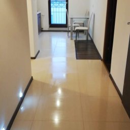 Tomasz Onyśko - Jasne, błyszczące płytki podłogowe w korytarzu z oświetleniem punktowym przy podłodze. W tle widoczne okno balkonowe i minimalistyczny zestaw stół z krzesłami.