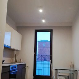 Tomasz Onyśko - Jasna kuchnia z ciemnymi szafkami i widokiem na ceglaną wieżę przez balkon. Minimalistyczny stół z krzesłami. Podłoga z połyskiem odbija światło.