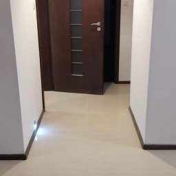 Tomasz Onyśko - Jasne płytki podłogowe w korytarzu z dwiema drewnianymi ościeżnicami i drzwiami z przeszkleniami. Białe ściany i sufit z lampą.