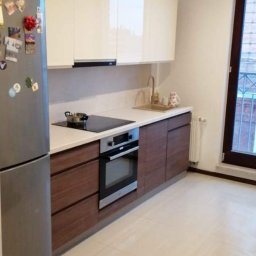 Tomasz Onyśko - Nowoczesna kuchnia z brązowymi szafkami dolnymi i białymi górnymi, jasnymi płytkami podłogowymi i srebrną lodówką z magnesami. Widok na okno z kratą.