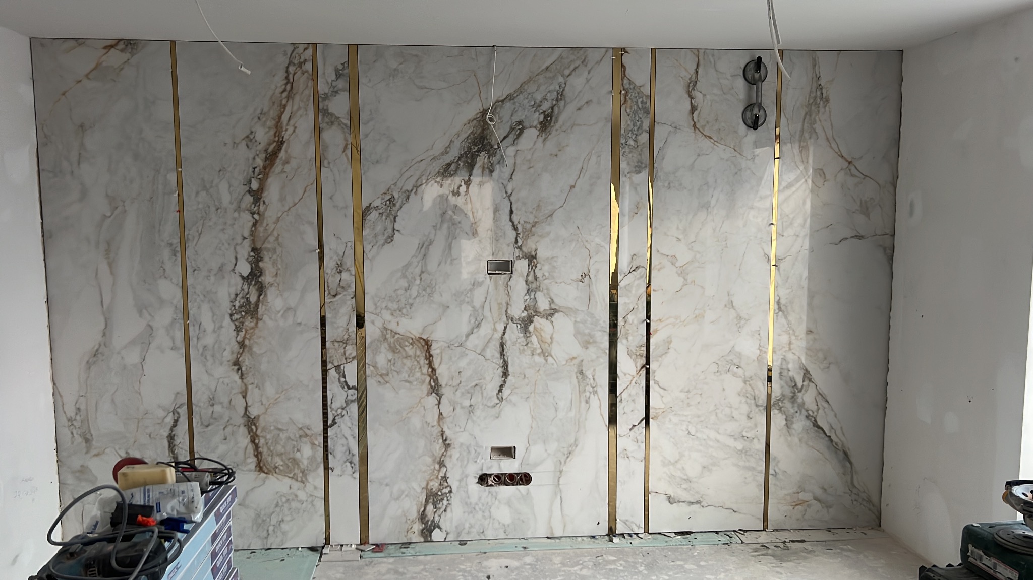 Ściana wyłożona wielkoformatowymi płytami imitującymi marmur, z cienkimi, pionowymi, złotymi listwami dekoracyjnymi, widoczne puszki elektryczne i narzędzia glazurnicze w dolnej części kadru.